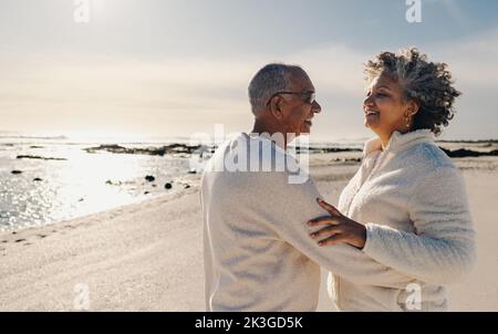 Mature couple smiling and dancing together on beach sand. Happy elderly couple having a good time next to the ocean. Cheerful senior couple enjoying t Stock Photo