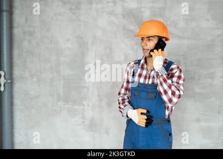 Muslim young man or builder in helmet with clipboard calling on smartphone over gray concrete wall background. repair, building, construction, busines Stock Photo