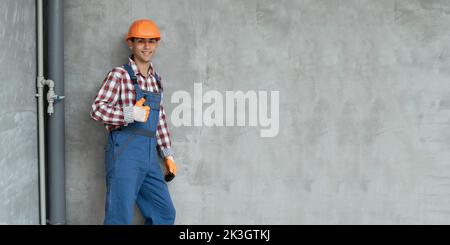 happy smiling male worker or builder in yellow helmet and overall showing thumb up over gray background. Banner. profession, construction and building Stock Photo