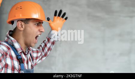 Screaming builder in helmet. Young man construction worker in hard hat crazy and mad shouting and yelling with aggressive expression and arms raised. Stock Photo