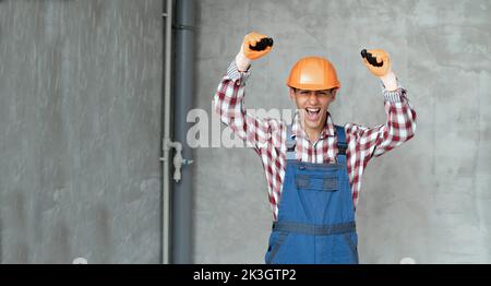 Photo of a happy positive young man builder in helmet posing isolated over gray wall background using mobile phone. profession, construction and build Stock Photo