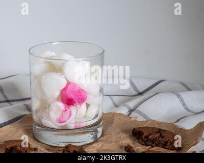 White Ice cubes made from milk in heart shape with one red heart in glass. Space for text. Cocoa cookies with chocolate on paper on a white background Stock Photo