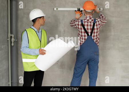 A young civil engineer with blueprints in his hands checks the work of a builder doing repairs in the house. Builder in a protective helmet measures t Stock Photo