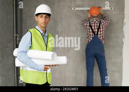 Architect man with blueprints and a working builder in the background against the wall, teamwork at a construction site. repair, building and remodeli Stock Photo