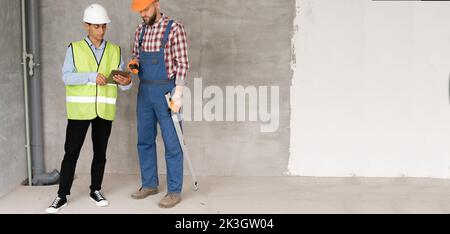 Two male builder and engineer in safety helmets are working with a tablet at a construction site, discussing blueprints for home renovation. building, Stock Photo