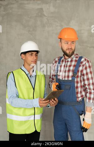 Working in team. Two young and cheerful builders in protective helmets are using digital tablet and working while standing on gray background. Copy sp Stock Photo