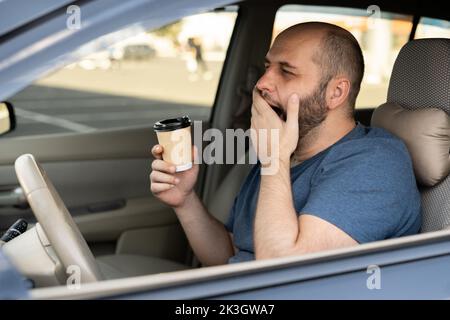 Adult handsome man feeling tired and yawning while driving a car. Driver having coffee early morning in avto Stock Photo