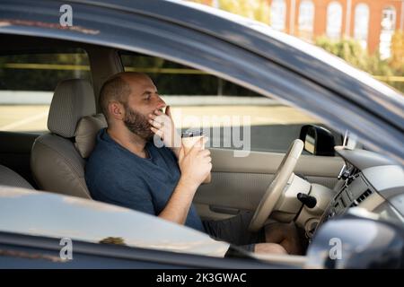 Tired man yawning on the front seat of the car holding coffee to go. Transportation sleep deprivation accident Stock Photo