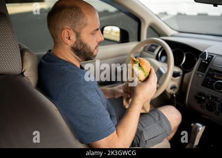 Man eating an hamburger while driving car. Hungry driver driving his car while eating food in the traffic Stock Photo