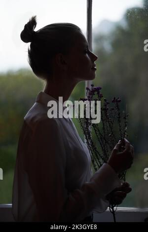 Let your imagination take flight. A young woman standing by a window holding flowers. Stock Photo