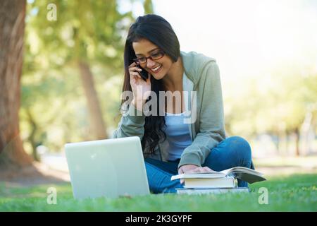Meet me in the park for a study session. A young student speaking on her cell while sitting in the park with a laptop and testbooks. Stock Photo