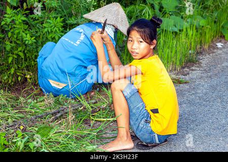 Vietnamese girl sitting by roadside with worker trimming crops, Hai Phong, Vietnam Stock Photo