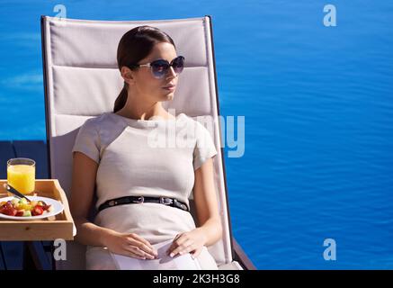 Relaxing while on holiday. A posh young woman sitting on a deck chair by the poolside. Stock Photo