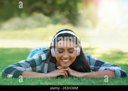 Take the music with you. A young woman listening to music over her headphones while lying in the park. Stock Photo