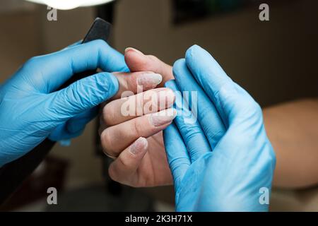 Close-up image manicurist in blue gloves holds client hand, examines  nail plate after removing old gel polish coating. Female client receives manicur Stock Photo