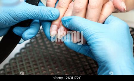Close-up image manicurist in blue gloves holds client hand, compares the length and shape of woman customer nails after removing old material gel poli Stock Photo