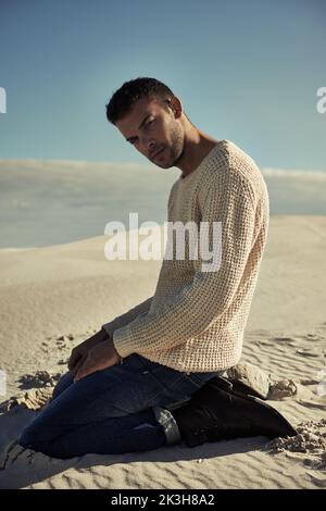 Man vs. desert. Handsome man kneeling on the desert sand in trendy clothing. Stock Photo