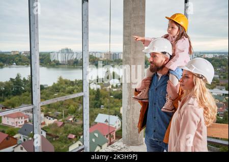 Daughter sitting on father's shoulders and pointing at something at construction site. Family with child future homeowners observing apartment building under construction. Stock Photo