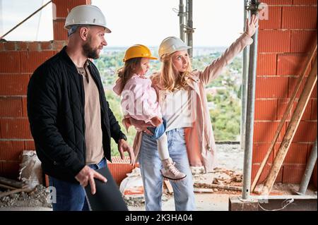 Woman in safety helmet holding daughter and discussing construction with builder. Female future homeowner talking with worker in apartment building under construction. Stock Photo