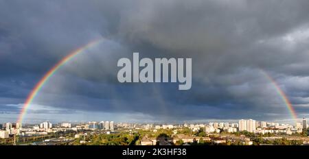 Large rainbow semicircle in the cloudy sky over the city. City landscape after the rain in a light blur. Stock Photo