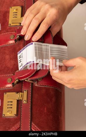 Enngland, UK. 2022. Airline luggage destination identity tag being attached to a dark red leather suitcase handle by womans hands. Stock Photo