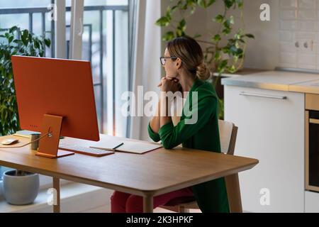 Distracted from study or job serious woman sitting at table with computer, looking to window at home Stock Photo