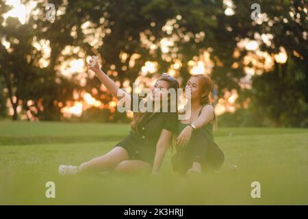 Two young Asian women smile for the camera as they take a selfie together. Both are sat on the grass in a park on a beautiful summer day at sunset. Stock Photo