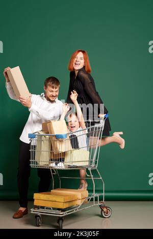 Joyful family, mother, father and son with shopping bags and boxes ride on shop cart isolated on green background. Concept of sales, black friday Stock Photo