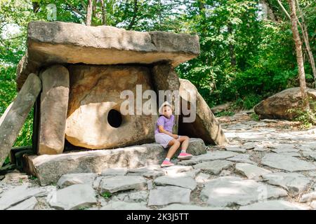 Little girl sit near ancient dolmen in green park on sunny day. Tourists look at natural sights on summer vacation. Single chamber megalithic tomb Stock Photo