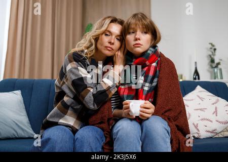 Sad girl with flu in a blanket and with a cup of tea and her mother sitting on the sofa in the living room. Pretty mature woman hugged her beloved dau Stock Photo