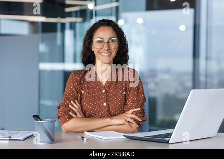 Portrait of successful young businesswoman, Hispanic woman working inside modern office building smiling and looking at camera sitting at desk with laptop, female worker with curly hair and glasses Stock Photo