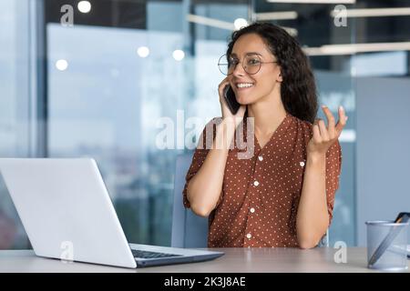 Young successful hispanic businesswoman working inside modern office, female employee cheerfully talking to colleagues on phone and smiling, using laptop while sitting at desk. Stock Photo