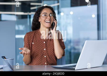 Young successful hispanic businesswoman working inside modern office, female employee cheerfully talking to colleagues on phone and smiling, using laptop while sitting at desk. Stock Photo
