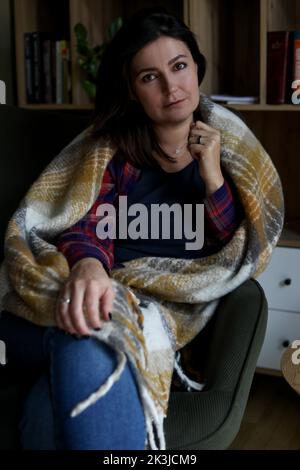 Vertical mature thoughtful, calm brunette woman, covered by blanket, looking at camera and sit in armchair in library Stock Photo