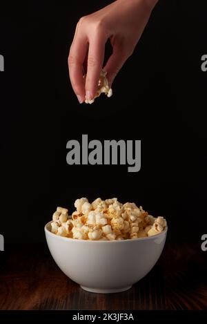 Woman’s hand taking popcorn from full white porcelain bowl on dark rustic wooden table and black background Stock Photo