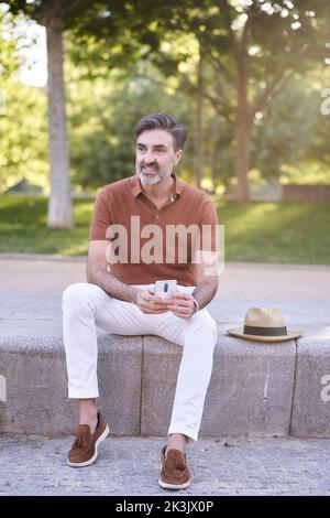Man looking away and relaxing while using a mobile phone outdoors in a park. Technology concept. Stock Photo