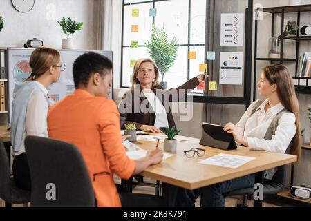 Senior blond financier woman pointing on board with paper sticks during speech on conference with multiracial female colleagues. Competent economists discussing company incomes at office room. Stock Photo