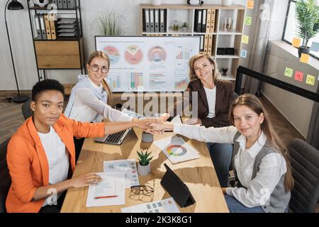 International team of competent female brokers holding their hands together in circle after profitable cooperation at office room. Modern technology for work on desk. Teamwork concept. Stock Photo