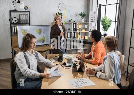 Senior woman showing on glass board financial statistic of company for multicultural female partners at modern office. Business people using modern technology for work. Conference process. Stock Photo