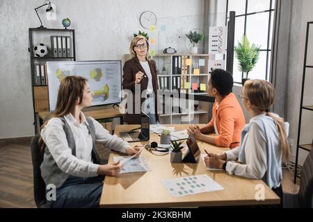 Senior woman showing on glass board financial statistic of company for multicultural female partners at modern office. Business people using modern technology for work. Conference process. Stock Photo