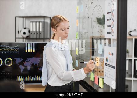Young female speaker at formal wear writing on transparent board during conference at office room. Company worker presenting financial report during business meeting. Stock Photo
