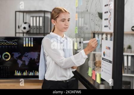 Young female speaker at formal wear writing on transparent board during conference at office room. Company worker presenting financial report during business meeting. Stock Photo