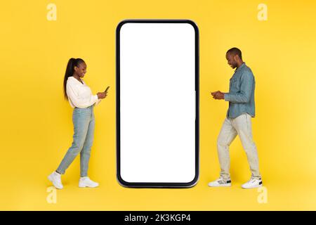 African Couple Using Phones Posing Near Large Cellphone, Yellow Background Stock Photo