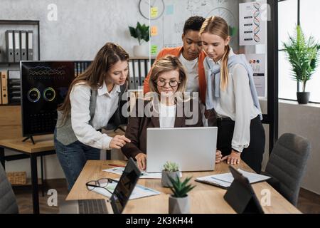 Group of confident high-skilled multiracial business women, standing at the table and discussing business strategy, while senior aged lady working on project on laptop computer. Stock Photo
