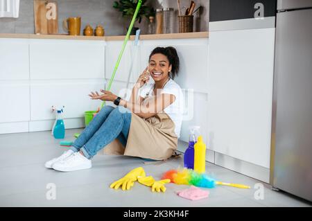 Happy millennial black woman sits on floor with mop, cleaning supplies, calls by phone in kitchen interior Stock Photo