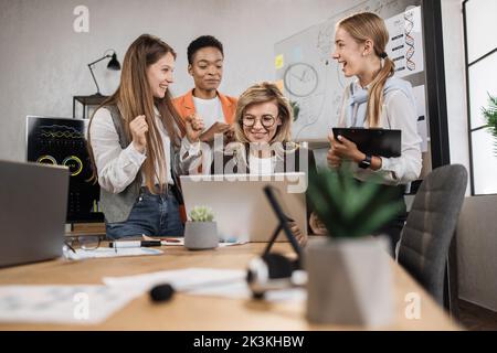 Close up of confident multiethnic businesswomen with senior female leader, working together in office, using laptop and tablet pc, discussing business strategy or new startup Stock Photo