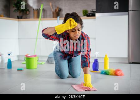Tired unhappy young black lady washes floor with cleaning supplies, wipes sweat from forehead Stock Photo