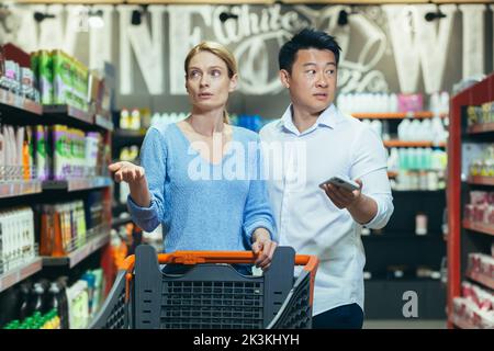 Confused young international couple, woman and Asian man walking between rows with grocery cart and phone in handsin supermarket. They spread their hands, looking for goods. Stock Photo