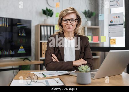 Beautiful senior woman with blond hair looking at camera while sitting at desk with papers and modern laptop. Business lady in stylish formal suit posing at own office Stock Photo