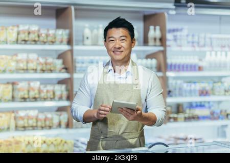Portrait of a young handsome Asian man, shop owner, a supermarket worker. Standing in an apron in the dairy department of the store with a notebook in his hands, looking at the camera, smiling. Stock Photo